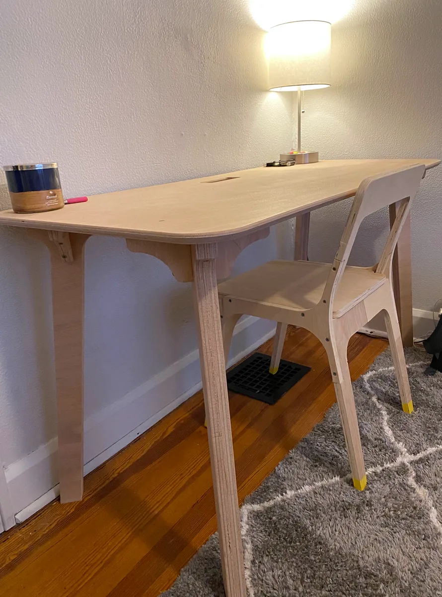 A plywood desk and chair stand against a wall beneath a glowing table lamp.