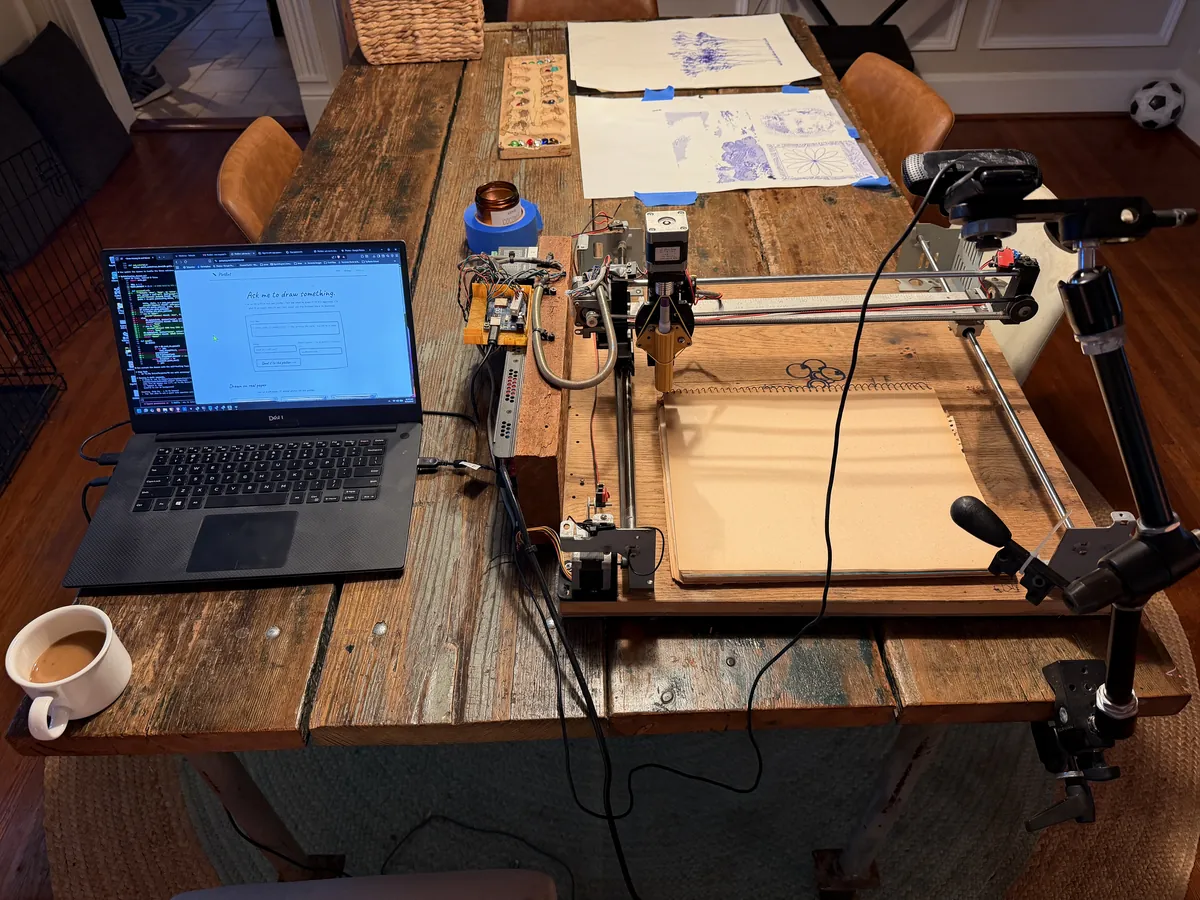 The pen plotter on a wooden dining table: a laptop, the Arduino controller, the gantry over a sketchbook, and a webcam on an arm above the bed