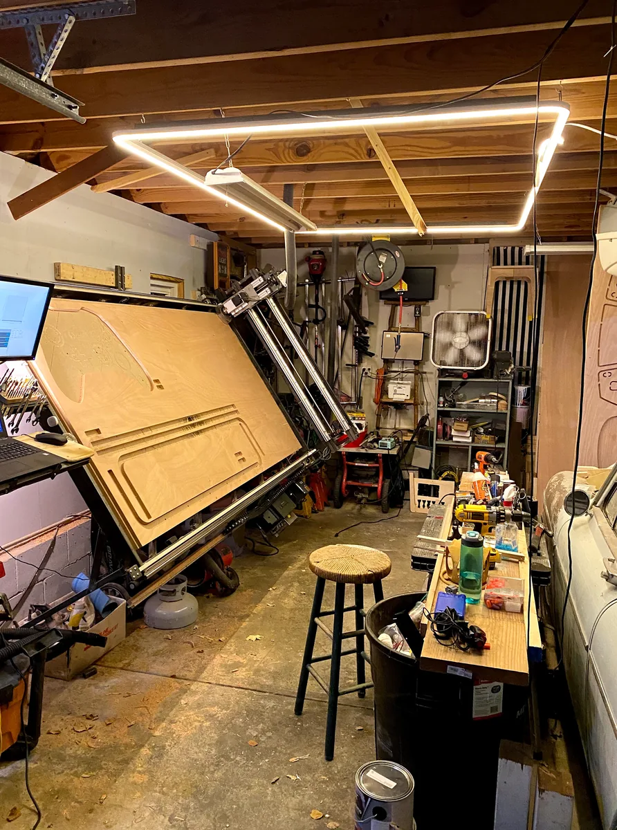 A tilted CNC router, workbench, and tools fill a workshop beneath exposed ceiling joists.